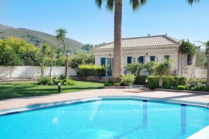 Una piscina con borde de azulejos, rodeada por un jardín ajardinado con palmeras y arbustos, y un edificio con techo de tejas al fondo.