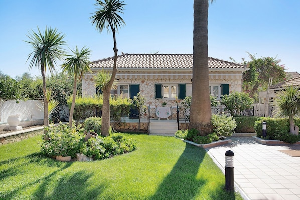 Una casa con techo de tejas, fachada de piedra y un jardín bien cuidado con palmeras y arbustos.
