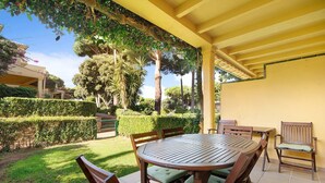 Une terrasse couverte avec une table ronde en bois et des chaises, entourée de haies et d’arbres soigneusement taillés.