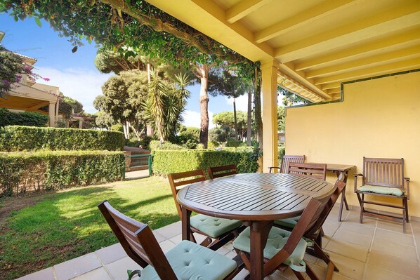 Une terrasse couverte avec une table ronde en bois et des chaises, entourée de haies et d’arbres soigneusement taillés.
