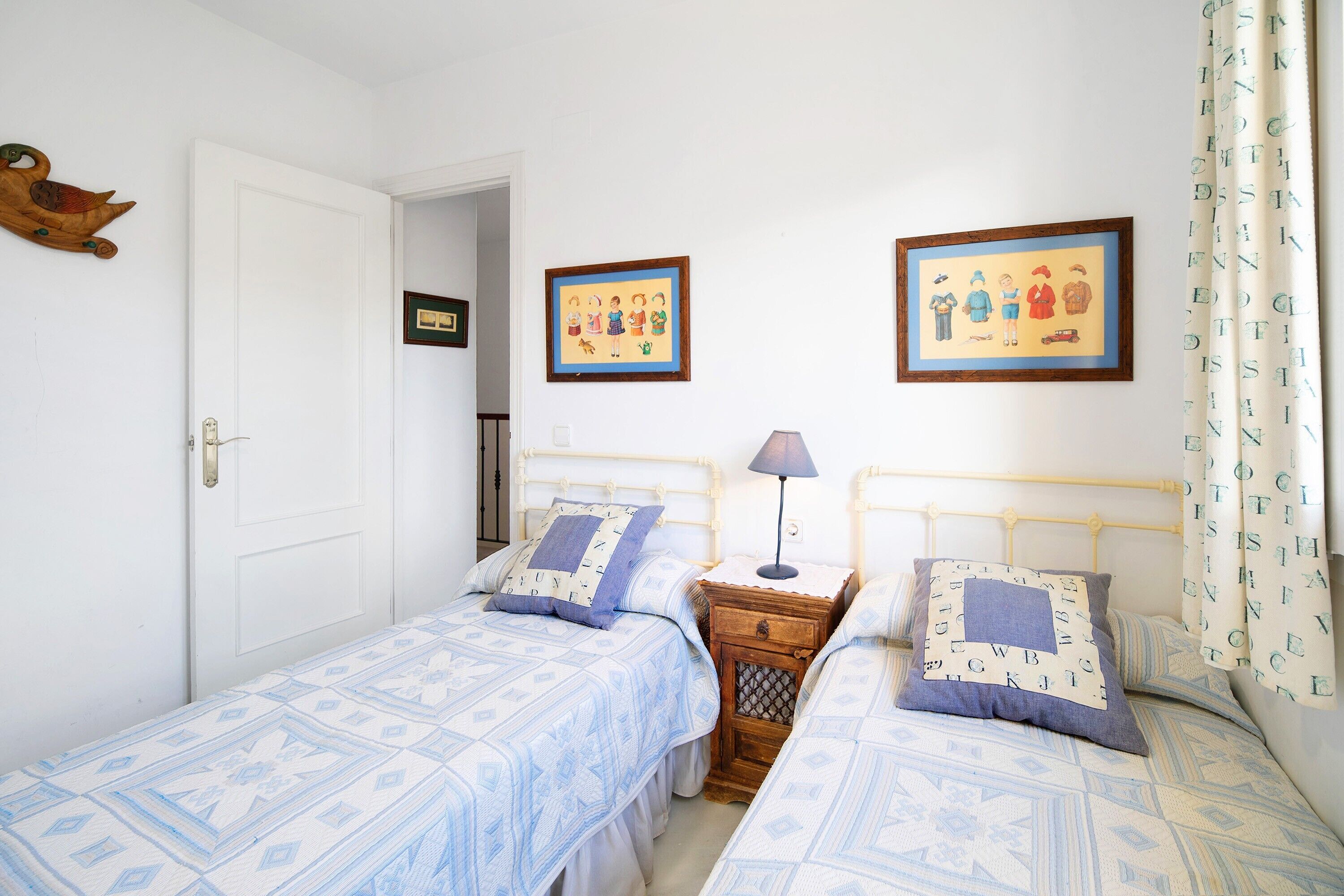 Quarto com duas camas, uma mesa de cabeceira de madeira, uma luminária e duas fotografias emolduradas na parede.