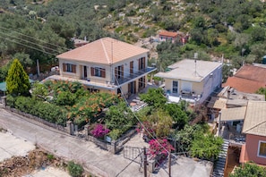 A two-story house with a balcony, surrounded by greenery and a paved driveway.