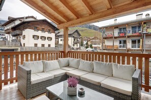 Ein überdachter Außenbereich mit Sitzgelegenheiten, einer Korbsessel-Couch und einem Glastisch, mit Blick auf Berge und traditionelle Gebäude.