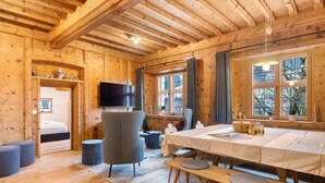 Interior de una cabaña de madera con una mesa de comedor, sillas, un banco, un televisor y ventanas amplias.