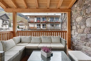 Un balcon abrité avec un plafond en bois, un canapé en osier agrémenté de coussins, une table basse en verre et un mur en pierre.