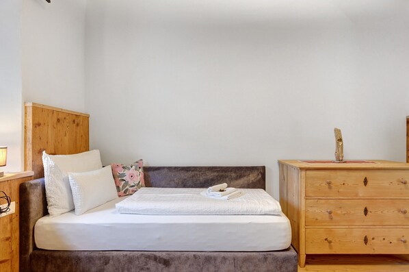 Ein Bett mit weißer Bettwäsche, ein Holz-Kopfteil, ein Nachttisch mit Lampe und eine Kommode aus Holz mit Schubladen.