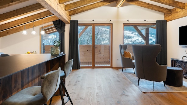 A modern interior with exposed wooden beams, a dining area with a long table and chairs, a seating area with a sofa and armchair, and large windows offering a view of the outdoors. - Apartment 'Alpin Chalet Samer Fürstenblick' with Mountain View, Terrace and Wi-Fi (Sarntal)