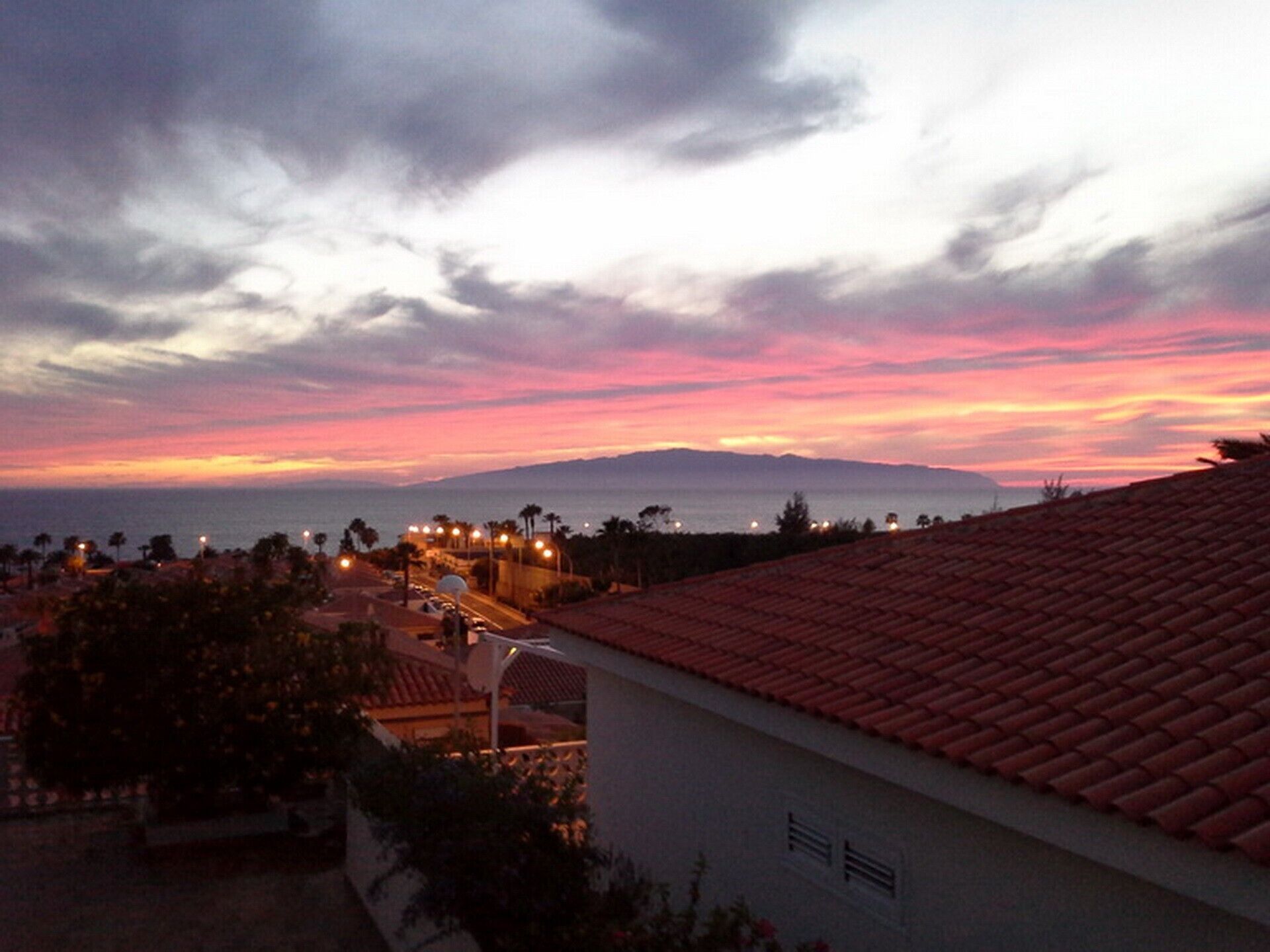 A coastal view with a sunset, buildings with red-tiled roofs, and a distant mountain range.