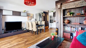 Ein modernes Wohnzimmer mit Flachbildfernseher, Essbereich, Bücherregalen und einer roten Pendelleuchte.