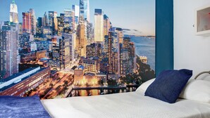 Ein Schlafzimmer mit Stadtbild-Wandgemälde, einem Bett mit weißer Bettwäsche und einem blauen Kopfkissen.