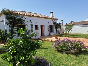 A white building with a tiled roof, a small garden with various plants, and a clear blue sky.