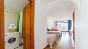 Zona de bugaderia amb rentadora, una porta de fusta i un passadís cap a un dormitori amb dos llits i una finestra.