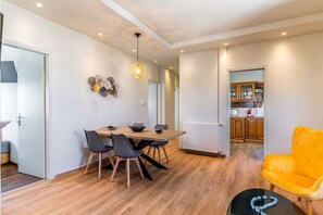 Un comedor moderno con una mesa y sillas de madera, una lámpara colgante y una cocina visible a través de un umbral abierto.