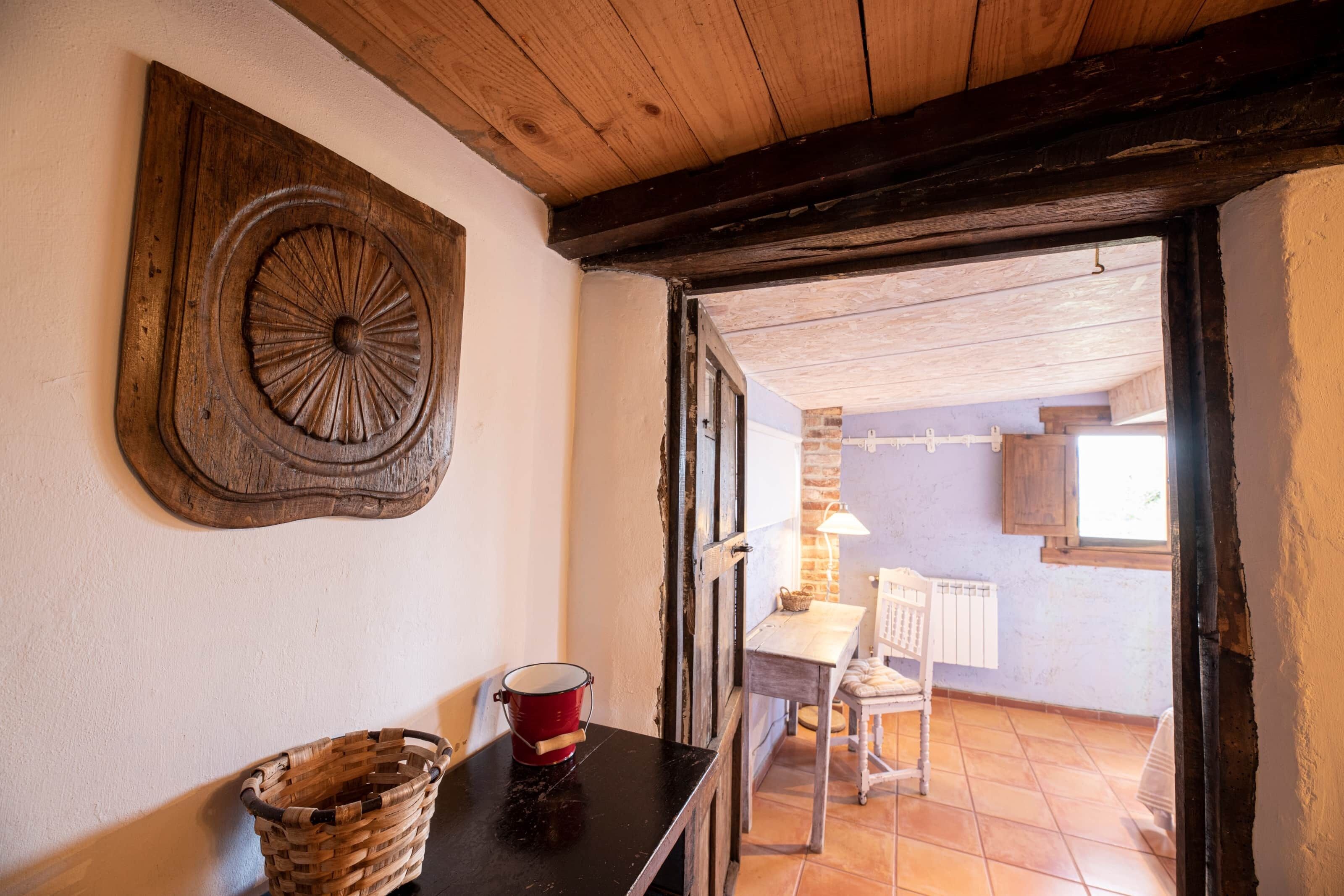 Ein rustikaler Raum mit Holzbalkendecke, einem an der Wand befestigten Wandkunstwerk, einem Tisch mit einem roten Eimer und Blick auf eine Steinwand und ein Fenster.