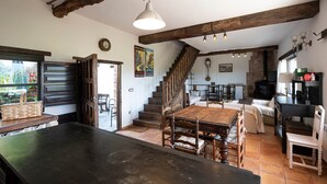 Un interno rustico con travi a vista in legno, un'area pranzo con un grande tavolo e sedie in legno, una scala che conduce a un altro livello e un angolo cottura con fornello e pensili.