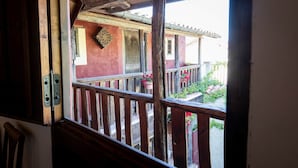 Un balcone in legno con ringhiera, vista su un edificio con pareti rosse e un elemento decorativo a parete, e piante in vaso con fiori.
