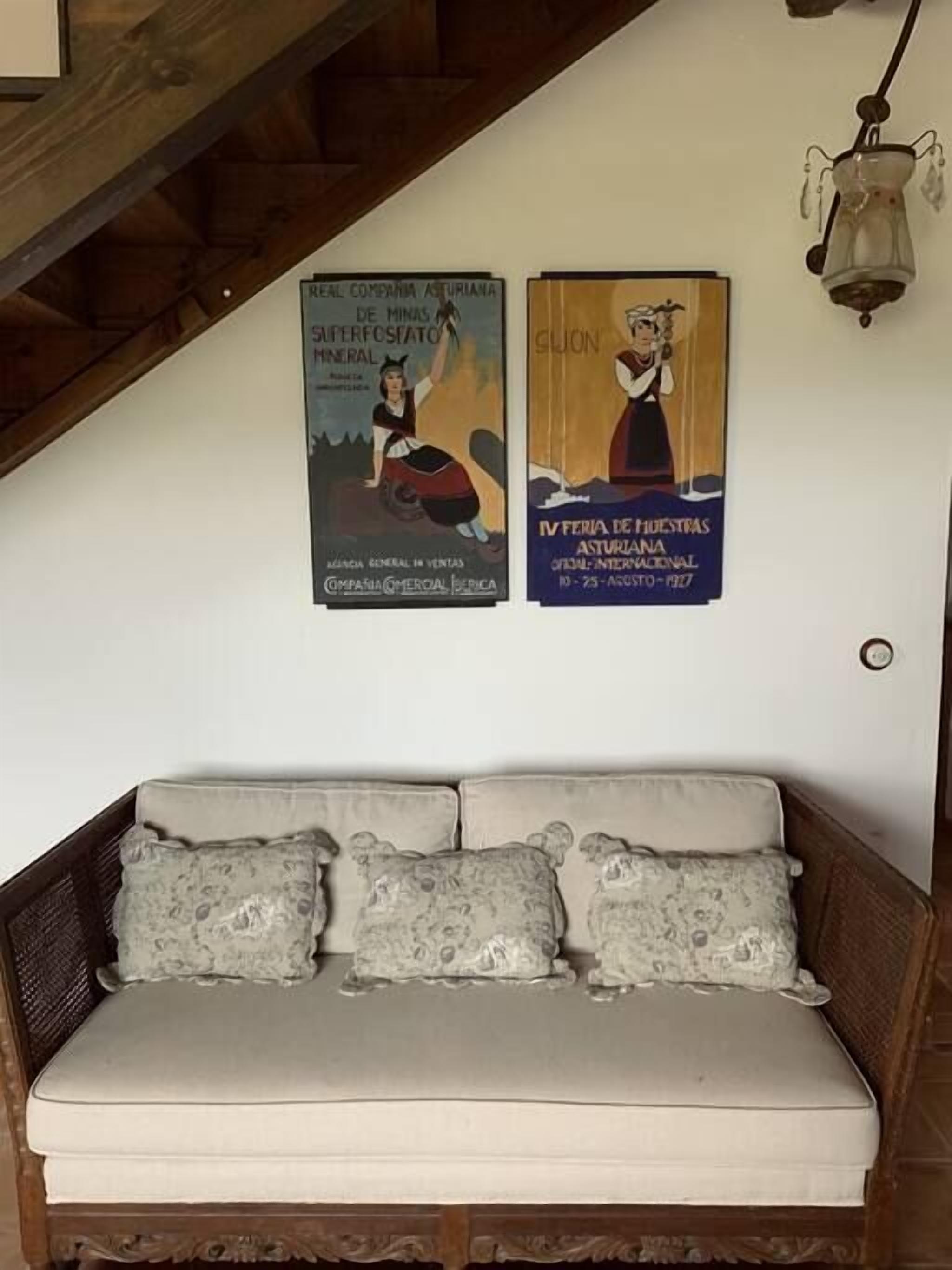 Ein Zimmer mit Holzboden, einer weißen Wand und einer Couch mit gemusterten Kissen. Zwei Poster sind an der Wand angebracht.