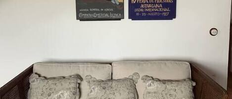 Ein Zimmer mit Holzboden, einer weißen Wand und einer Couch mit gemusterten Kissen. Zwei Poster sind an der Wand angebracht.