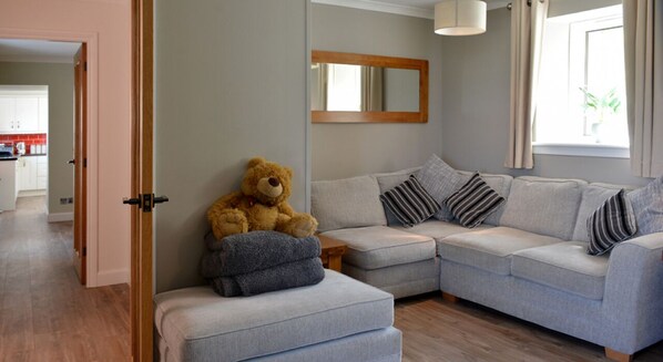 A living room with a large grey sectional sofa, a small ottoman, and a teddy bear. There is a wooden door leading to a kitchen, a mirror on the wall, and a window with curtains.