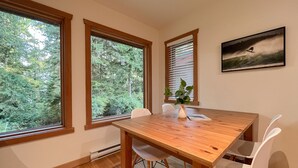 A dining area with a wooden table, white chairs, a vase with a plant, and large windows with a view of trees. - Eagle's Nest Suite-Inlet, Forest Views, Marina (Tofino)