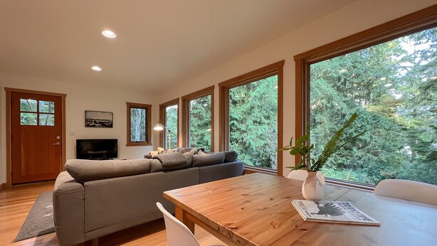 A modern living room with a wooden dining table, a grey sofa, and large windows offering a view of trees. - Eagle's Nest Suite-Inlet, Forest Views, Marina (Tofino)