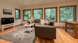 A modern living room with a sectional sofa, a flat-screen TV, a dining table, and large windows offering a view of trees. - Eagle's Nest Suite-Inlet, Forest Views, Marina (Tofino)