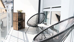 Un balcon avec deux chaises en fil de fer noir, une petite table ronde et une caisse en bois contenant des plantes.