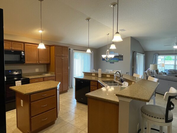 A modern kitchen with granite countertops, wooden cabinets, and a central island. There is a dining area with a table and chairs, and a living room with a sofa and a lamp. - Beautiful 3/2 Villa Walking distance to Brownwood w/BBQ & Golf Cart! (The Villages)