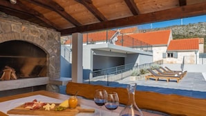 A stone fireplace, outdoor seating with sun loungers, and a view of a hillside with buildings.