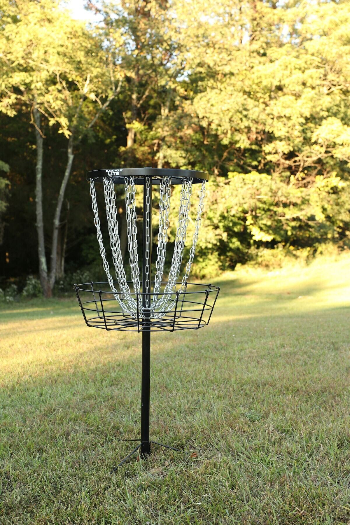 Een frisbee golfmand met een metalen frame en kettingen, gelegen op een grasveld met bomen op de achtergrond.
