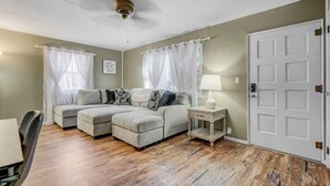 Ein Zimmer mit einem Ecksofa, einem kleinen Tisch mit Lampe, einer Tür und einem Deckenventilator.