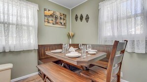Ein Esszimmer mit einem Holztisch, gedeckt für sechs Personen, an beiden Seiten flankiert von Fenstern mit weißen Spitzenvorhängen.