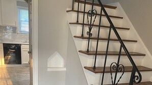A staircase with wooden steps and black metal railings, leading to a room with white walls and a kitchen area visible in the background. - Cozy Detroit home in the Center of Metro Detroit. (Detroit)