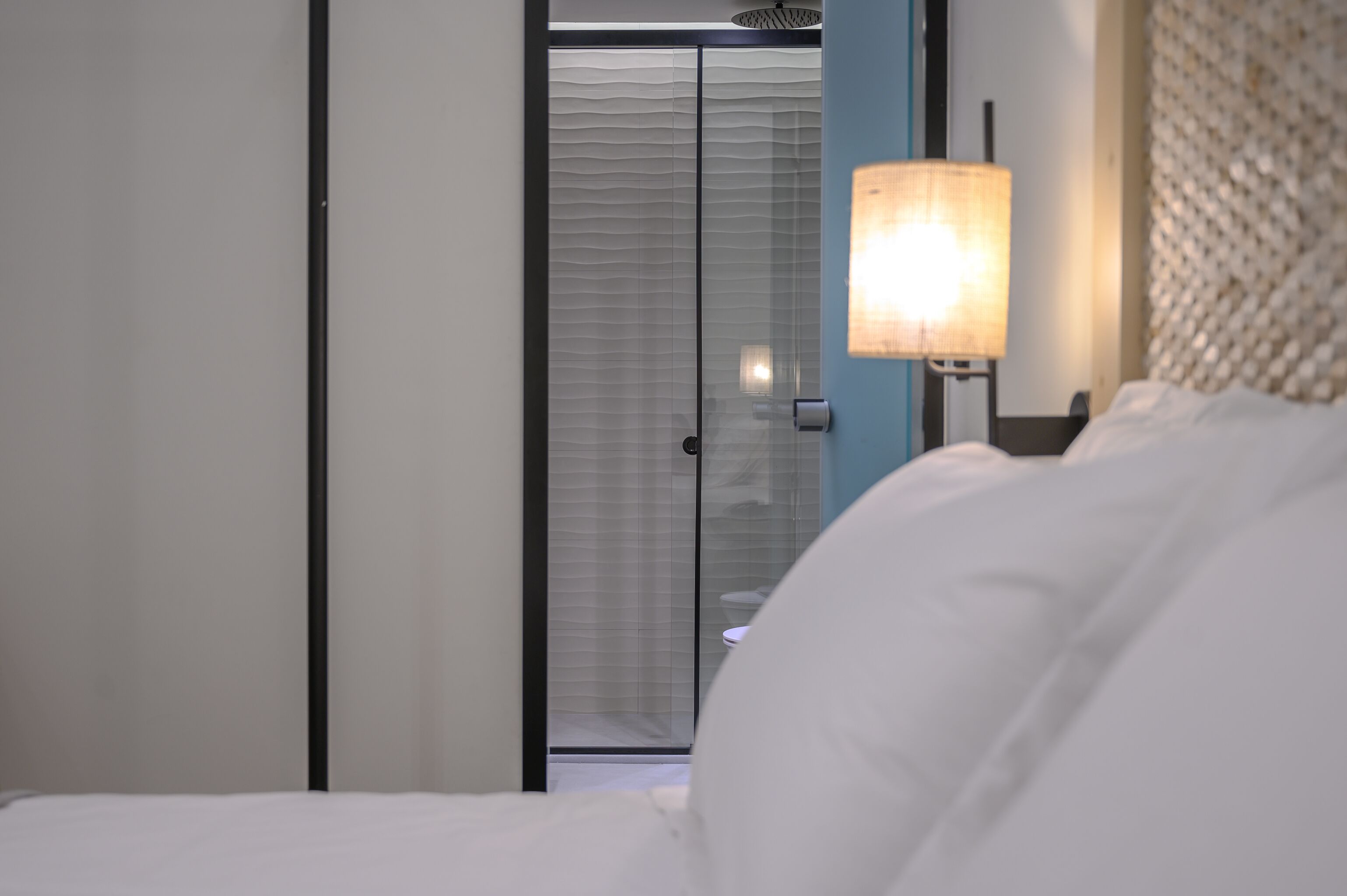 A hotel room with a bed, a nightstand with a lamp, a glass door leading to a bathroom, and a blue wall.