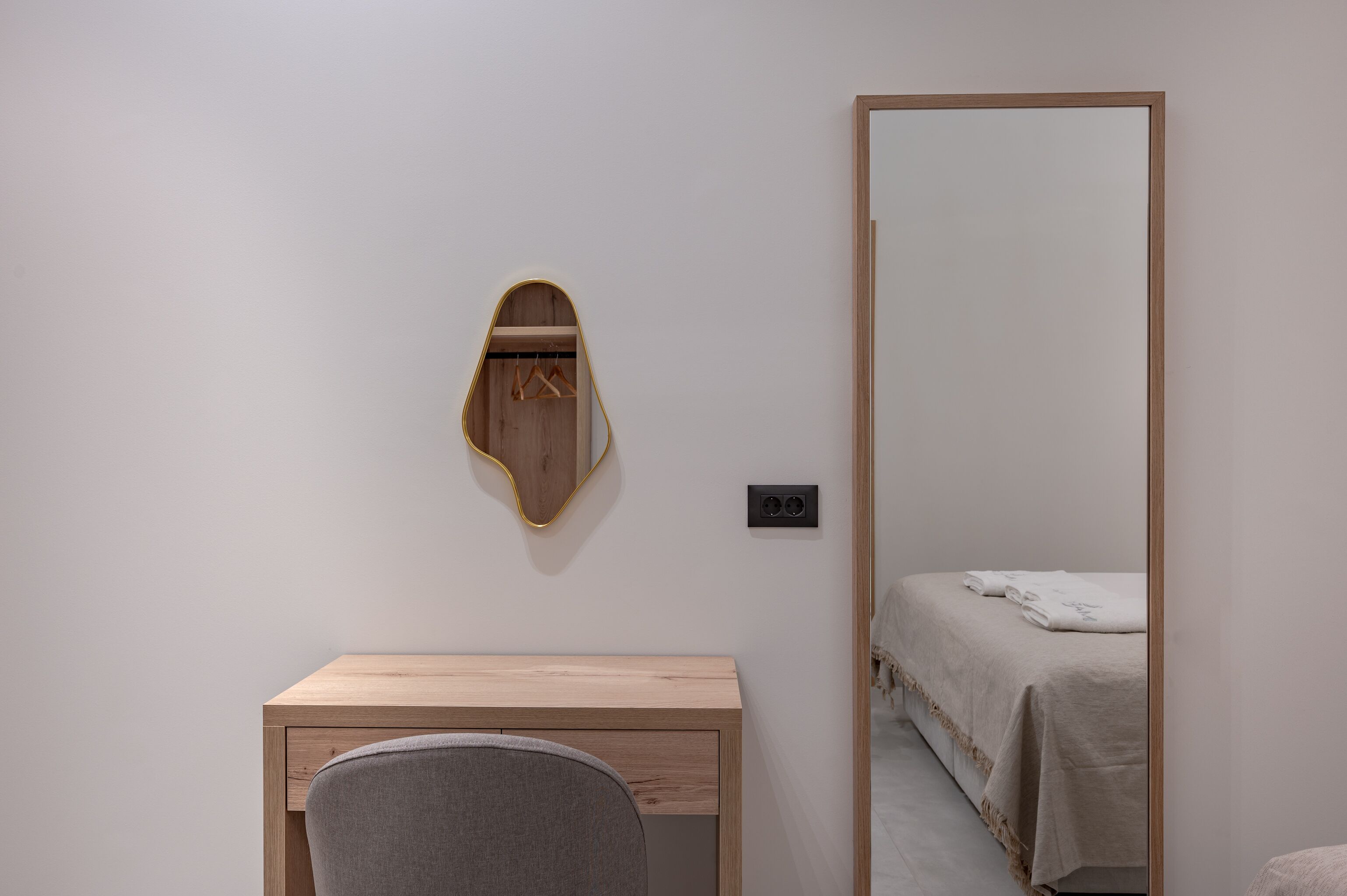 A minimalist room with a wooden desk, a chair, a bed with a white bedspread, and a large mirror with a wooden frame.