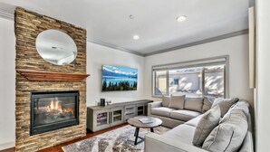 Una sala de estar moderna con chimenea de piedra, un televisor de pantalla plana grande, un sofá y una ventana con vistas a una casa.