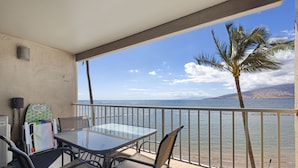 Un balcó amb taula i cadires de vidre, amb vista a una platja que té una palmera i un cel serè.