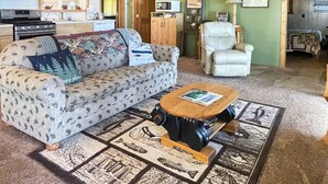 En hyggelig stue med sofa, lænestol, sofabord og et køkken, der ses gennem en åben dør.