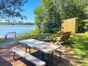 Un tavolo da picnic con panche, un braciere e una piccola struttura in legno accanto a un lago.