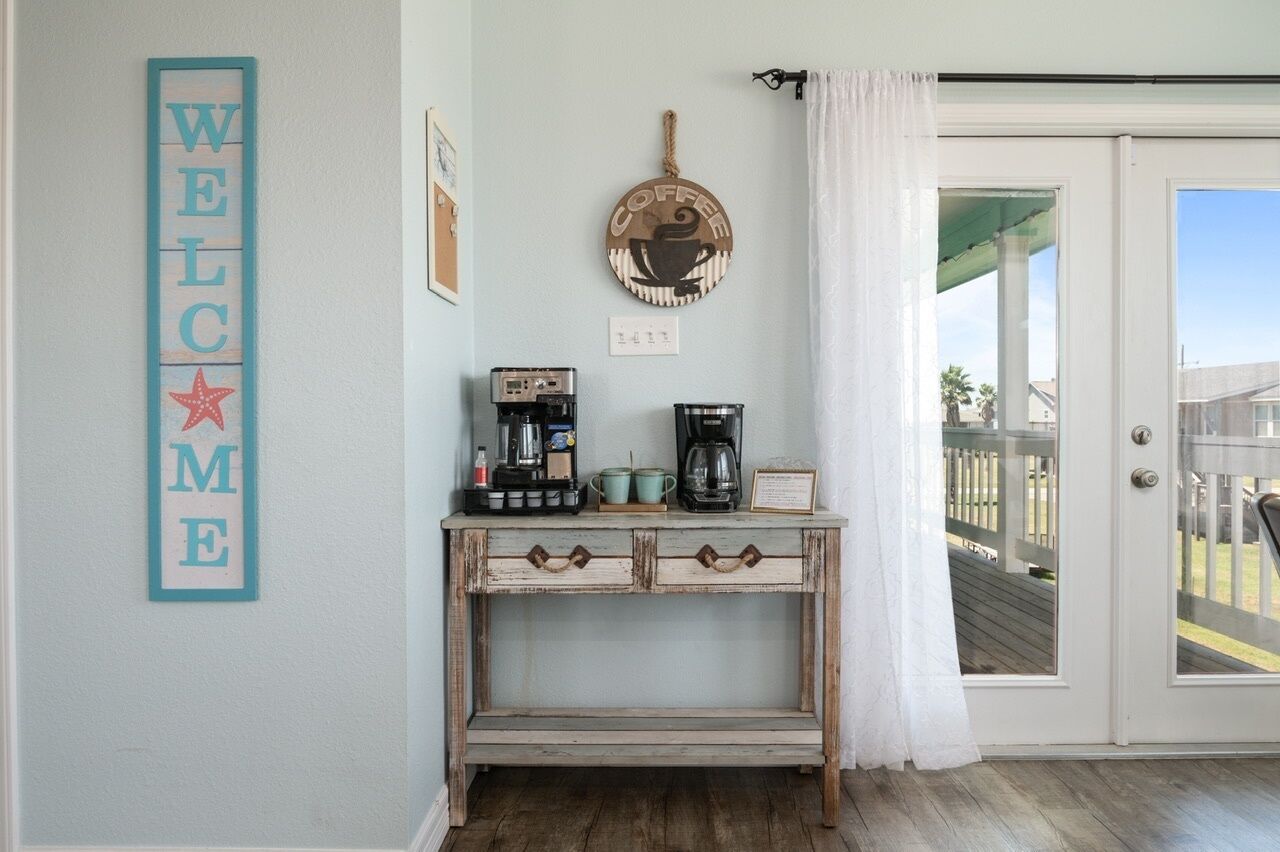 Una habitación con un cartel de 'WELCOME', una estación de café con cafetera, tazas y un letrero, y una puerta que da a un espacio exterior.