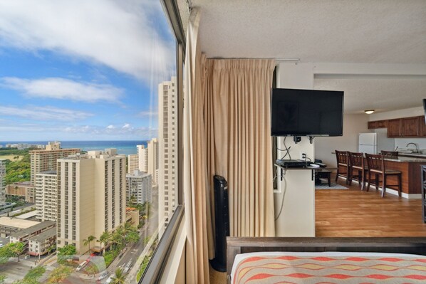 A hotel room with a large window, a flat-screen TV, a kitchenette, and a dining area with chairs. - Tower 1 Suite 2705 - QQS (Honolulu)