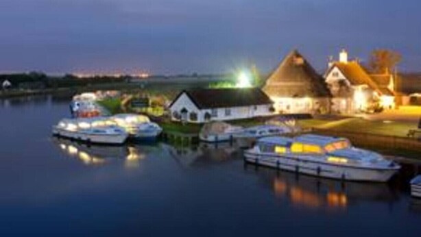 Ein Haus am Wasser mit angestaubten Booten, einem Reetdach und einem gut beleuchteten Bereich, der abendliche Aktivitäten andeutet.