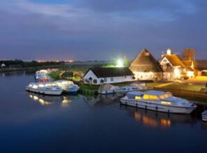 A waterfront property with boats docked, a thatched-roof house, and a well-lit area suggesting evening activities. - Stunning 2 Bed Georgian Apartment (North Walsham)