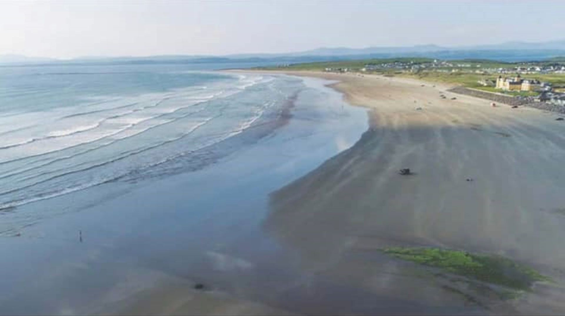 Vista aérea de una playa con orilla arenosa y un pequeño poblado al fondo.