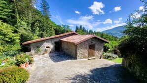 A stone cottage with a tiled roof, surrounded by lush greenery and mountains in the background. - Beautifully converted watermill with private pool and own waterfall! (San Romano in Garfagnana)