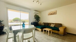 Ein Wohnzimmer mit Esstisch, Stühlen, einer Couch und einem kleinen Couchtisch. Ein großes Fenster bietet Blick auf Gebäude und Bäume.