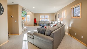 A spacious living room with a grey sectional sofa, a television, and a window with blinds.