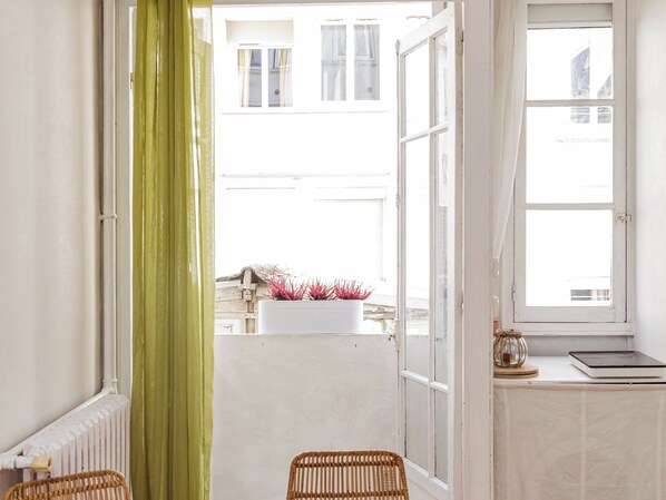 A room with a view of a building, a window with a curtain, a radiator, and a wicker chair.