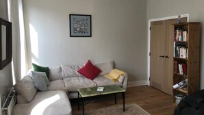 Una sala de estar con un sofá beige, una mesita de centro pequeña, una estantería y un cuadro enmarcado en la pared.
