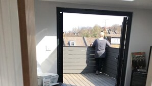 Una habitación con piso de madera, una mesita con una silla, una puerta corrediza que da a un espacio exterior donde hay una persona de pie y un cartel en la pared.